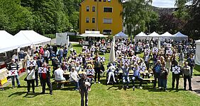 Viele Menschen sitzen auf Bierbänken und an Tischen auf einer Wiese. Einige halten Schilder mit einem roten X hoch.