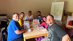 Sechs Menschen sitzen an einem kleinen Tisch zusammen. Hinter ihnen steht ein Flip-Chart.
