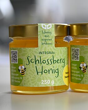 Zwei Gläser Honig stehen auf einem Tisch. Sie tragen ein Etikett mit dem Aufdruck Schlossberg-Honig.
