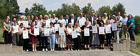 Eine große Gruppe von Menschen steht zum Gruppenbild zusammen. Alle halten ein Zertifikat in den Händen.