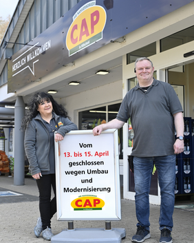 Zwei Mitarbeiter stehen vor dem CAP-Markt. Sie lehnen sich an ein Schild an, auf dem über die Schließung des Marktes für Umbauarbeiten informiert wird.