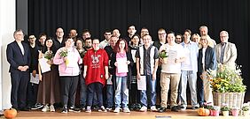 Mehr als 30 Menschen stehen auf einer Bühne. Einige halten ein Zertifikat oder Blumen in den Händen.