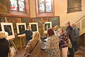 Eine Gruppe von Menschen steht am Altar einer Kirche. Auf dem Altar steht ein Kruzifix. Hinter dem Altar stehen mehrere Bilder auf Staffeleien.