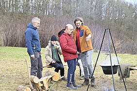Vier Menschen stehen im Freien an einer Feuerstelle. Über der Feuerstelle hängt ein Kessel mit dampfendem Inhalt an einem Dreibein.