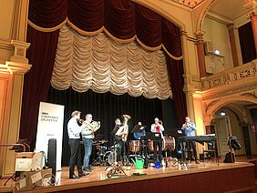 [Translate to Leichte Sprache:] Sechs Musiker stehen auf einer großen Bühne. Sie spielen Blechblasinstrumente und Schlaginstrumente.