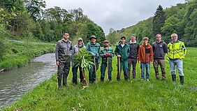 Eine Gruppe von Männern steht auf einer Wiese am Ufer eines Baches. Sie tragen Arbeitskleidung. Einer von ihnen hält einen Riesenbärenklau.