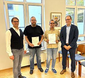 Vier Männer stehen nebeneinander und schauen in die Kamera. Die beiden mittleren halten ein Zeugnis in den Händen.