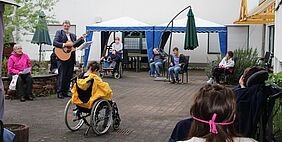 Ein Mann spielt unter freiem Himmel die Gitarre für ein Publikum. Einiger Rollstuhlfahrer sind darunter. 
