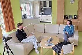Ein Mann und eine Frau sitzen auf einem großen Sofa. Sie unterhalten sich miteinander. Dabei filmt sie eine Kamera.