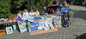 Eine Gruppe Menschen sitzt hinter einem Stand auf einem Kunstmarkt. Es sind viele Bilder zu sehen, die zum Verkauf angeboten werden.