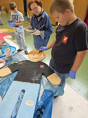Ein Schüler mal eine Holzfigur als Junge an, eine Frau im Malerkittel sieht zu