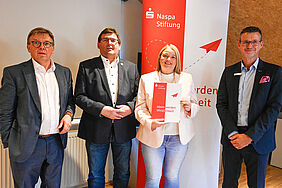 Vier Männer und eine Frau stehen vor einem Roll-up der Naspa-Stiftung.