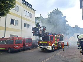 Feuerwehrfahrzeuge stehen vor einem mehrstöckigen Haus, aus dem Qualm dringt. Im Korb der Drehleiter befinden sich Menschen.