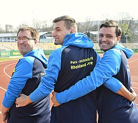 Drei junge Männer stehen mit dem Rücken zum Betrachter. Sie wenden ihre Köpfe um, sodass man ihre Gesichter sieht. Sie tragen Trainingsanzüge des BSV.