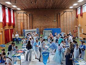 In einer Turnhalle sind viele Menschen in Malkitteln unterwegs und arbeiten an Tischen