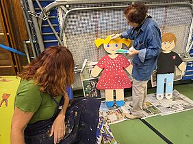 eine Frau bemalt kindliche Figuren aus Holz, eine andere Frau schaut zu