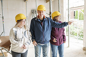 In einem Rohbau zeigt ein Mann zur Seit. Zwei Frauen schauen in diese Richtung. Sie tragen Bauarbeiterhelme. 