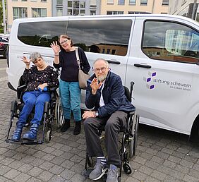 Drei Personen vor einem neuen weißen Auto und winken in die Kamera.