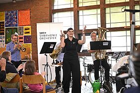 Eine Frau klatscht in die Hände zur Musik. Hinter ihr stehen Musiker und spielen auf ihren Blechblasinstrumenten.
