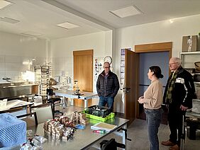 Eine Frau stellt zwei Männern die Geräte und Seifenblöcke in einem Arbeitsraum vor