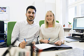 Ein Mann und eine Frau sitzen an einem Schreibtisch vor einem Order mit Papieren. 