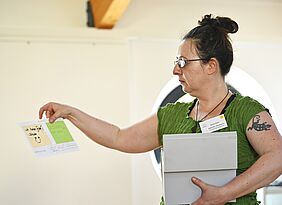 Eine Frau hält einen Kasten unter dem Arm, mit einer Hand hält sie eine beschriftete Karteikarte.