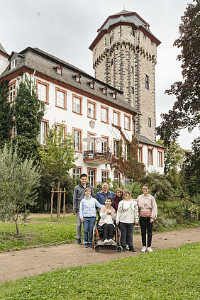 Eine Gruppe mit einer Rollstuhlfahrerin in einem Park vor einem Schloss. 