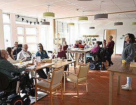 Klienten und Mitarbeitende sitzen und stehen an Tischen. Einige halten Zettel in den Höhe, auf denen die Spendensumme ablesbar ist.